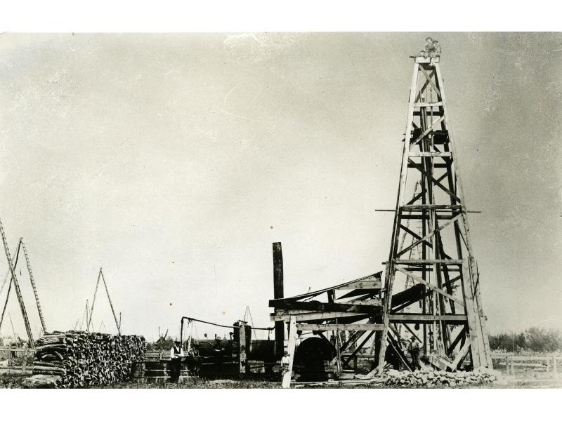 A wooden oil rig with  four legs and crossbeams moving up its height. There are oil derricks off to the left behind a stack of wood.