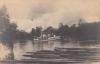 Front of postcard showing a white boat on a river. There is a pile of logs in the foreground and trees in the background.  