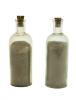 Two clear glass bottles filled with sand-coloured ash. The bottles are tall with a short spout and light brown corks in the tops.