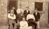A photo of eight men in front of a stone building with a wooden-framed door and window. Four are standing at the back, three are sitting in chairs, and one is sitting on the ground.