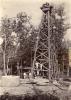 A photo of a group of International Drillers standing at the base and on the first support beam of an oil derrick. They are wearing white. There is a large cylinder and some logs in front of them and trees in the background.