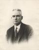 A photo of John Growder from the chest up. He is wearing a dark jacket and vest with a white shirt and dotted tie. He is clean-shaven with grey hair. 