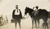 George Rawlings in a dark suit and riding boots. He is holding a pith helmet in one hand and the reins of a horse in the other. A man is standing behind the horse.  