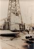 A photo of the bottom half of an oil rig being thatched. A group of men stand at the base. There are piles of casing on either side. 